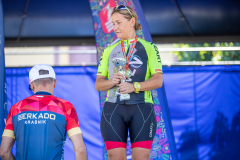 Kobieta w stroju kolarskim stoi na pierwszym miejscu podium i trzyma w rękach zdobyty puchar