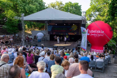 Widok na kilkadziesiąt osób siedzących na widowni umieszczonej dookoła małej sceny. Na scenie stoją dwie osoby. Po jednej stronie sceny stoi duży balon promocyjny z napisem Lubelskie Smakuj życie