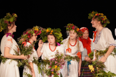 Kilka młodych dziewcząt wystawiających sztukę teatralną. Są ubrane w białe sukienki i maja na głowach wieńce z kwiatów