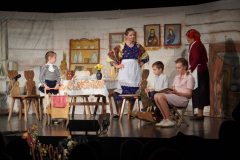 Ujęcie podczas przedstawienia teatralnego, dwie dorosłe kobiety i trójka dzieci w otoczeniu scenografii stylizowanej na wnętrze chłopskiej chaty