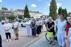 Kilkanaście osób, mieszkańcy miasta stoją na chodniku i przyglądają się uroczystościom. Za nimi w tle widać miejskie budynki, trwa letnia słoneczna pogoda