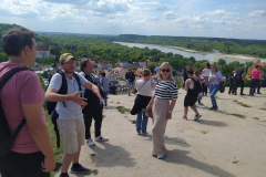 Kilkanaście osób w letnich strojach stoi na piaszczystym wzgórzu, pod nimi rozciąga się widok na zieloną dolinę z płynącą przez niąrzeką