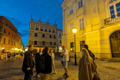 Kilka osób stoi nocą na placu Starego Miasta i ogląda znajdujące się na nim kamienice