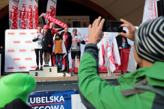 Na podium stoi kilka osób które prezentują zdobyte medale, jedna z nich trzyma szalik z napisem Polska. Na pierwszym planie stoi osoba, która robi zdjecie stojącym na podium