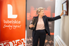 Eliza Kunz podczas prezentowania wystawy fotograficznej. Wskazuje na obramowane zdjęcie i opowiada.