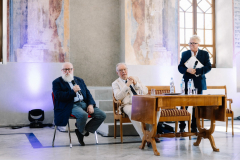 Prof. Jerzy Bralczyk siedzi na krześle w sali przypominającej pałacowe wnętrze obok niego znajduje się dwóch innych mężczyzn