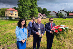 Sabina Bober, Jarosław Stawiarski, Józef Rudy stoją na cmentarzu żydowskim, trzymają wiązankę kwiatów oraz znicz. Za nimi widać rosnącą trawę a w dali znajdują się budynki mieszkalne