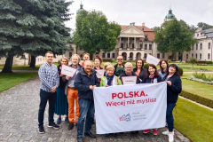 Kilkanaście osób pozuje do zdjęcia stojąc w dwurzędzie. Dwie osoby z przodu grupy trzymają baner na którym jest napisane: Polska, więcej niż myślisz Polska Organizacja Turystyczna. W tle widok na kompleks pałacowy.