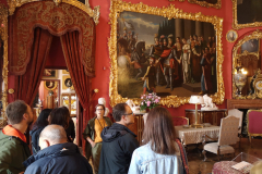 Grupa kilku osób zwiedza wnętrze pałacu. W  pomieszczeniu widać ściany zapełnione obrazami i przejście  do dalszych pokoi, które jest ozdobione kotarą.