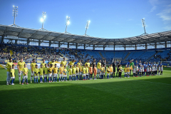 Zawodnicy obu drużyn piłakrskich stoją w jednym rzedzie na murawie na stadionie.