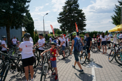 Kilkanaście osób, uczestników wyścigu w strojach sportowych, stoi przy swoich rowerach na otwartym placu.