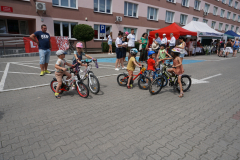 Kilkoro małych dzieci na swoich rowerkach stoją na placu. W tle widać dorosłych oraz rozstawione namioty promocyjne