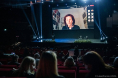 Sala operowa wypełniona ludźmi, na scenie widać ogromny ekran na którym wyświetla się kobieta łącząca się zdalnie na konferencję