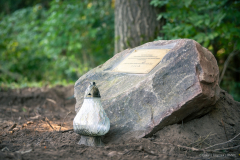 Widok na leżący w lesie obelisk z tablicą pamiątkową na nim i stojącym  palącym się zniczem