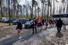 Grupa młodzieży szkolnej idzie w kolumnie przez las w poczcie sztandarowym, Wszyscy mają założone przez ramię biało-czerwone szarfy a jeden z chłopców niesie sztandar
