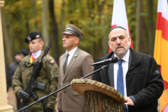 Naczelny Rabin Polski stoi przy mikrofonie ustawionym na zewnątrz i udziela wypowiedzi, za nim w tle widać dwóch wojskowych w mundurach i ustawione dwie flagi, polską i województwa lubelskiego