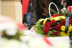 Widok na dłoń kobiecą, kobieta układa pod pomnikiem biały kamień jako symbol pamięci, na pierwszym planie widać ułożoną pod pomnikiem wiązankę z kwiatami