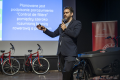 Prelegent Olivier Schneider przemawiający spacerując po scenie. W tle seria rowerów wraz ze specyficznym rowerem z dużym koszem doczepionym przed kierownicą, oraz prezentacja widniejąca na rzutniku.