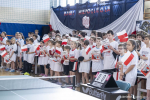 Kilkadziesiąt dzieci znajdujących się na sali gimnastycznej, wszyscy ubrani w jednakowe koszulki, trzymają w rękach papierowe flagi Polski i machają nimi