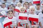 Widok na kilkanaścioro dzieci w wieku wczesnoszkolnym ubrane w jednakowe koszulki, wszystkie trzymają papierowe flagi Polski