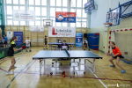 Dwa stoły do ping ponga przy których gra czwórka zawodników w wieku szkolnym. Na sali gimnastycznej jest powieszony baner z napisel Lubelskie Smakuj życie