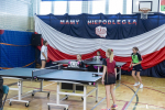 Cztery zawodniczki w wieku szkolnym grają w ping ponga przy  dwóch ustawionych stołach. Na drabinkach w sali gimnastycznej wisi dekoracja z biało czerwona flaga, i napisem mamy niepodległą