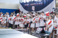 Kilkadziesiąt dzieci znajdujących się na sali gimnastycznej, wszyscy ubrani w jednakowe koszulki, trzymają w rękach papierowe flagi Polski i machają nimi