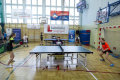Dwa stoły do ping ponga przy których gra czwórka zawodników w wieku szkolnym. Na sali gimnastycznej jest powieszony baner z napisel Lubelskie Smakuj życie
