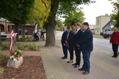 Uczestnicy wydarzenia stojący przed nowo odsłoniętą tablicą pamięci. Wśród zebranych znajduje się, między innymi, Wójt Gminy Roman Adamczyk. Tablica usytuowana jest na kamiennym pomniku przed budynkiem urzędu. W najbliższym otoczeniu można zauważyć liczne drzewa. Uczestnicy wydarzenia stojący przed nowo odsłoniętą tablicą pamięci. Wśród zebranych znajduje się, między innymi, Wójt Gminy Roman Adamczyk. Tablica usytuowana jest na kamiennym pomniku przed budynkiem urzędu. W najbliższym otoczeniu można zauważyć liczne drzewa.