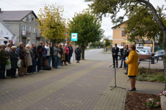 Liczna grupa uczestników wydarzenia, w szerokim przedziale wiekowym, stoi w rządku ze złożonymi rękoma na chodniku przed budynkiem urzędu. Przed nimi, kobieta wygłasza przemowę do stojącego mikrofonu. Liczna grupa uczestników wydarzenia, w szerokim przedziale wiekowym, stoi w rządku ze złożonymi rękoma na chodniku przed budynkiem urzędu. Przed nimi, kobieta wygłasza przemowę do stojącego mikrofonu.