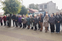 Liczna grupa uczestników wydarzenia, w szerokim przedziale wiekowym, stoi w rządku ze złożonymi rękoma na chodniku przed budynkiem urzędu. Jeden ze starszych mężczyzn wśród uczestników trzyma bukiet białych kwiatów. Liczna grupa uczestników wydarzenia, w szerokim przedziale wiekowym, stoi w rządku ze złożonymi rękoma na chodniku przed budynkiem urzędu. Jeden ze starszych mężczyzn wśród uczestników trzyma bukiet białych kwiatów.