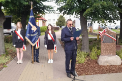 Wójt Gminy Roman Adamczyk odczytuje tekst z folderu trzymanego w dłoniach. Po jego lewej stronie znajduje się tablica pamięci owinięta biało-czerwoną szarfą. Za nim stoi trójka młodych ludzi ubranych na galowo i owiniętych podobnymi szarfami ze sztandarem Szkoły Podstawowej imienia Bohaterów Westerplatte w Michowie.
