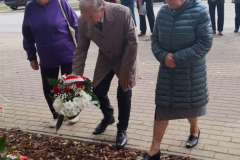 Trójka starszych wiekiem uczestników wydarzenia składająca bukiet kwiatów przy odsłoniętej tablicy pamięci. Trójka starszych wiekiem uczestników wydarzenia składająca bukiet kwiatów przy odsłoniętej tablicy pamięci.