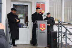 Ksiądz Biskup Mieczysław Cisło przemawiający z mównicy do zebranych na wydarzeniu. Po jego obu stronach stoi dwóch starszych mężczyzn - w tym Symcha Keller, natomiast za nim, jak i na przedzie mównicy, widoczny jest herb Gminy Firlej.