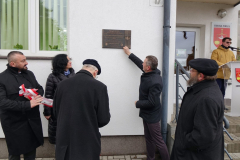 Uczestnicy wydarzenia otaczający odsłoniętą tablicę. W tle, osoba przemawia stojąc za mównicą.