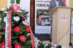 Członek Zarządu Marcin Szewczak stoi przy mównicy ustawionej przed budynkiem w letni dzień. Na pierwszym planie widać ustawione wieńce