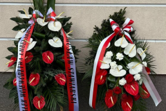 Dwa wieńce kwiatowe oraz dwa znicze ustawione pod nowo odsłoniętą tablicą pamięci. Kwiaty, oraz szarfy które owe wieńce ozdabiają, są w kolorach polskiej flagi.