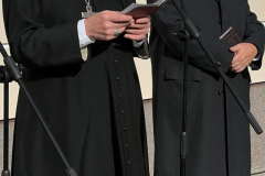 Biskup Mieczysław Cisło oraz Rabin Symcha Keller stojący z pochylonymi głowami przed budynkiem. Ksiądz biskup odczytuje do mikrofonu tekst z książeczki którą trzyma w dłoniach.