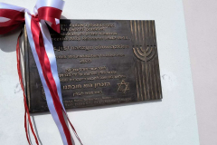 Tablica pamięci ozdobiona biało-czerwoną kokardą. Na tablicy widnieją upamiętniające napisy zarówno w języku polskim jak i hebrajskim. Kokarda przysłania częściowo treść tablicy. Tablica pamięci ozdobiona biało-czerwoną kokardą. Na tablicy widnieją upamiętniające napisy zarówno w języku polskim jak i hebrajskim. Kokarda przysłania częściowo treść tablicy.