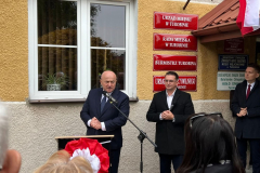 Marszałek Województwa Lubelskiego Jarosław Stawiarski przemawiający przy mównicy podczas odsłonięcia tablicy pamięci. Za nim stoi Burmistrz Andrzej Kozina oraz jeszcze jeden mężczyzna.
