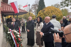 Burmistrz Andrzej Kozina przemawiający do mikrofonu, jednocześnie spoglądając na odsłoniętą tablicę. Tuż obok niego, bezpośrednio pod tablicą znajduje się wieniec z biało-czerwonymi kwiatami oraz szarfą. W otaczającym tablicę tłumie, na przedzie, stoją Marszałek Jarosław Stawiarski, Profesor Sabina Bober oraz Biskup Mieczysław Cisło. W pobliżu widoczne są polskie flagi.