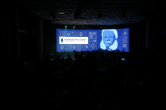 Widok na zaciemnioną salę oraz ekran na którym wyświetla się slajd, na którym widać podobiznę Antoniego Patka oraz napis w języku angielskim Gala passion for watches