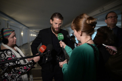 Sebastian Fabjański, aktor udziela wypowiedzi do mikrofonu, stoi w towarzystwie dziennikarzy, na zdjęciu znajdują się 4 osoby