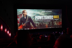 ekran w sali kinowej wyświetlający grafikę do filmu i napis Powstaniec 1863 na pierwszym planie widok na kilka pierwszych rzędów publiczności