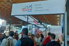 Stoisko promocyjne województwa lubelskiego dookoła którego przechodzi kilka osób, nad stoiskiem znajduje się baner z grafiką gór i lasu oraz napis anglojęzyczny polish tourism organisation