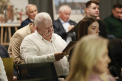 Widok na około dziesięć osób siedzących na sali konferencyjnej, obraz skupia sie na mężczyźnie czytającym konspekt