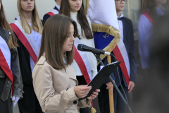 Młoda kobieta stoi przy mikrofonie i trzyma kartkę z której czyta w tle za nią widać poczet sztandarowy