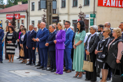 Około dwudziestu osób stoi wspólnie na placu na zewnątrz podczas letniej pogody.