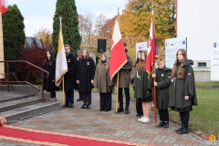 Młodzież z hufca harcerskiego stoi w poczcie sztandarowym. Około dziesięciorga dzieci stoi przed schodami prowadzącycmi do budynku, troje z nich trzyma sztandary