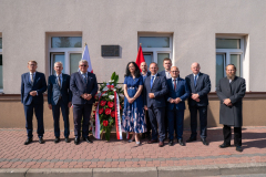 Wspólne zdjęcie uczestników odsłonięcia tablicy, przed budynkiem w miejscu na którym powieszona jest tablica stoi około dziesięciu osób, głównie przedstawicieli samorządu i lokalnych władz, pomiędzy nimi znajduje się wieniec